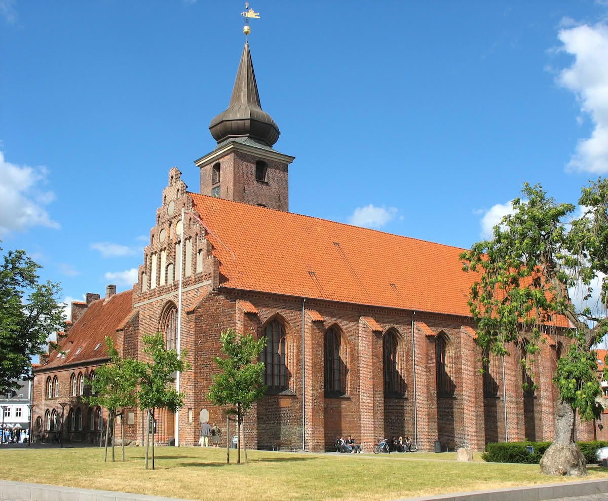 Klosterkirken — red-brick Gothic church in Nykøbing Falster, with a copper spire and summer trees